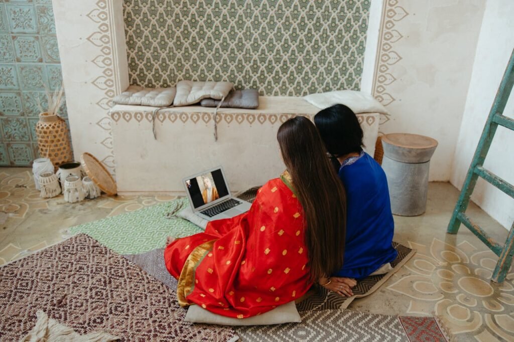 Two women in traditional clothing watch a laptop, sitting on patterned carpets indoors.